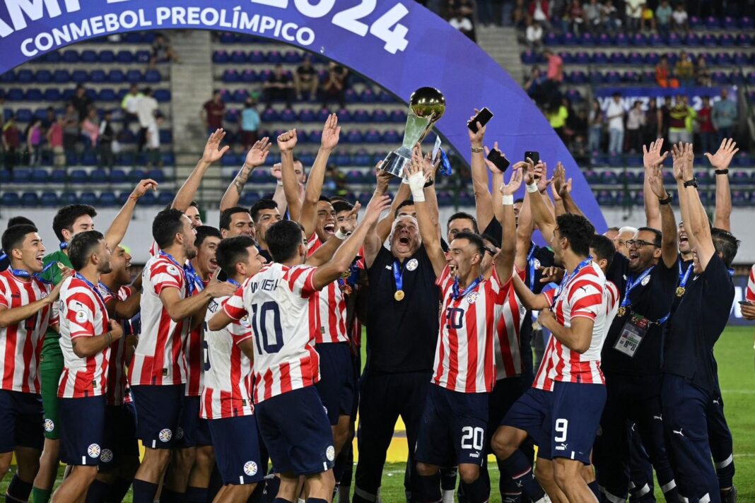 Jugadores de la selección paraguaya de fútbol levantan el trofeo de campeón del preolímpico. Detrás se visualiza un cartel que dice 